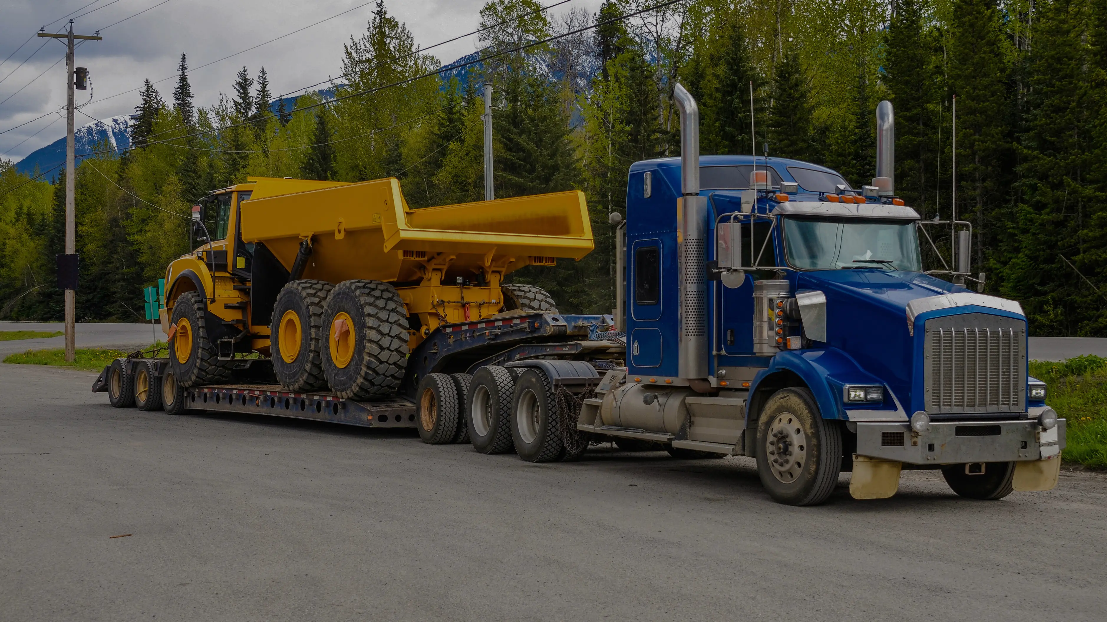 Blue truck transporting a yellow dump truck on a trailer, on a road surrounded by forests with mountains in the background