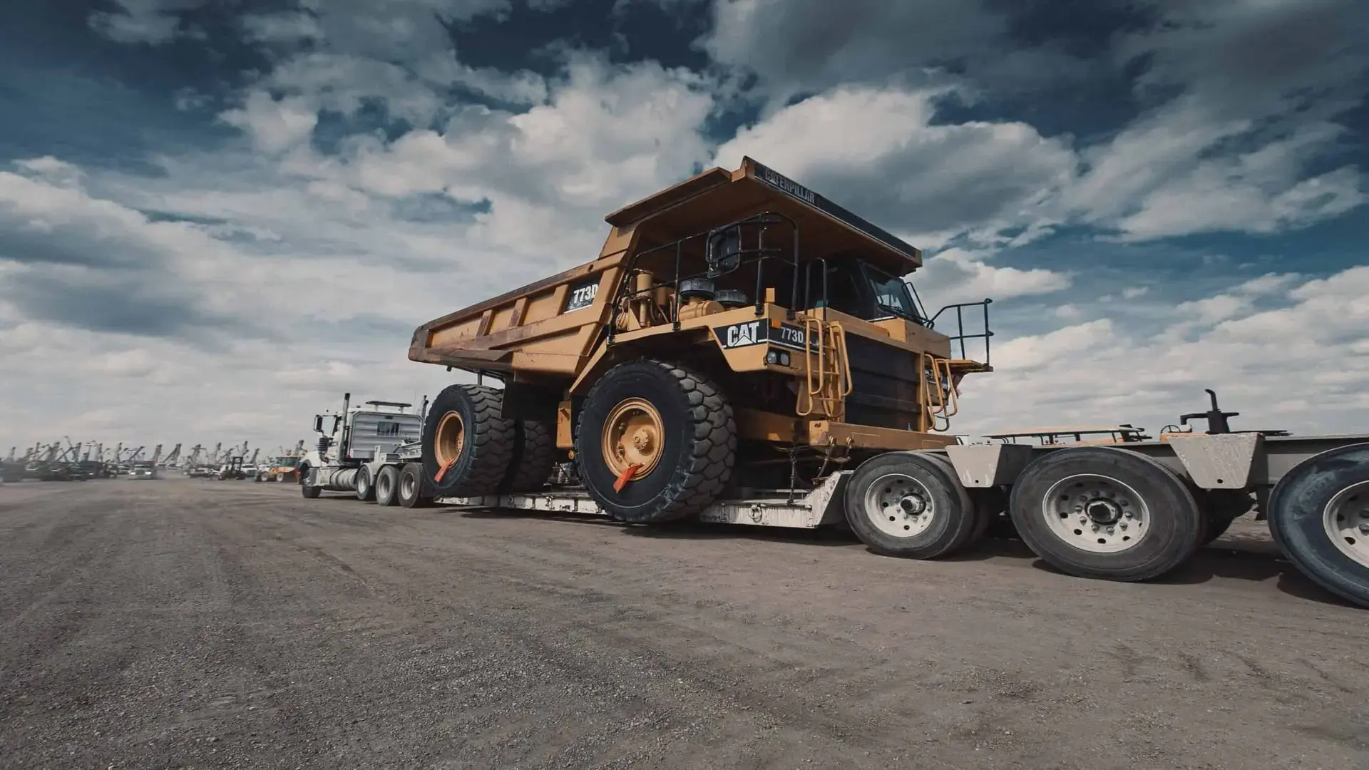 Transport truck carrying a large yellow dump truck on an open terrain under a cloudy sky