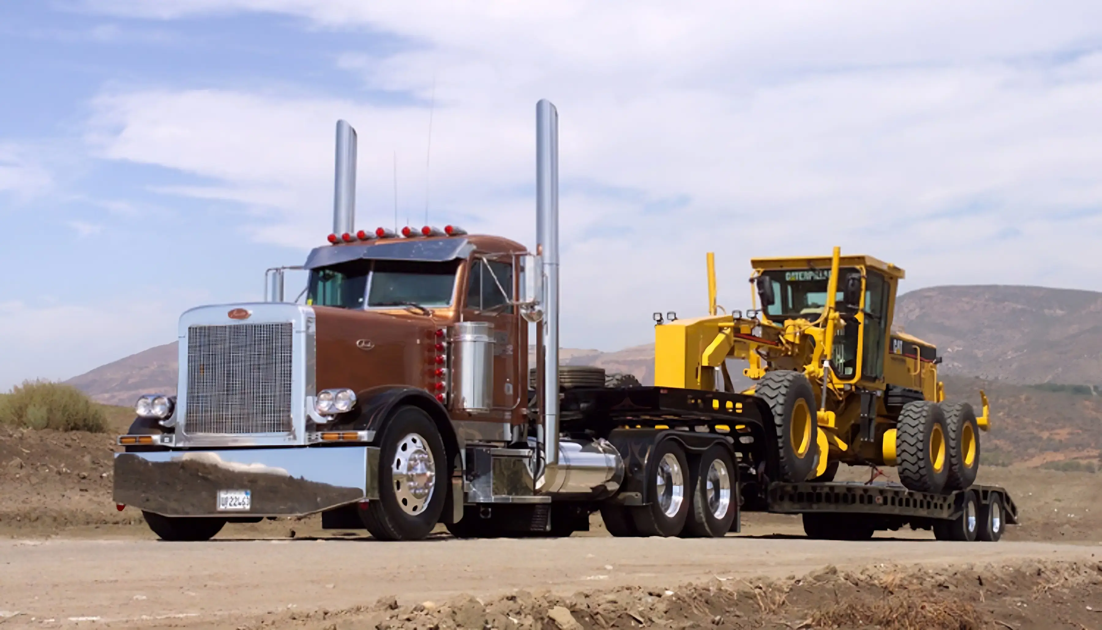 Brown truck transporting a yellow front loader on a trailer, set against a mountainous landscape with a clear sky